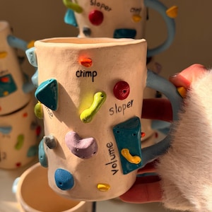 Puede incluir: Tazas de cerámica color crema con asas azules y presas de escalada coloridas. Las tazas tienen las palabras "crimp", "sloper" y "volume" escritas. Las presas de escalada son de varias formas y colores, incluyendo azul, verde y rojo.