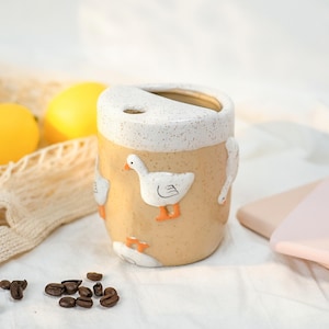 May include: A ceramic tumbler with a speckled tan body and a white speckled rim. The exterior features embossed white goose figures with orange beaks and feet. The top has a small circular opening and a larger slanted opening for drinking.