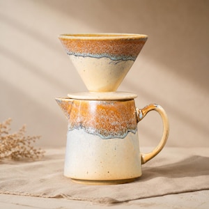 Op de afbeelding: Een keramische pour-over koffiezetapparaat set. De set bevat een crèmekleurige karaf met een handvat en een bijpassende kegelvormige druppelaar. De bovenkant van de karaf en de druppelaar hebben een bruin en wit glazuur. De set is ontworpen voor het zetten van koffie.