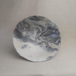 Puede incluir: Un plato de cerámica hecho a mano con una forma orgánica única. El plato presenta un diseño veteado en tonos grises y blancos, creando un patrón abstracto y fluido. El borde tiene un borde sutil e irregular, lo que aumenta su atractivo artesanal.