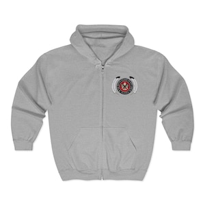 May include: A light gray zip-up hoodie with a hood and front pockets. The hoodie features a circular emblem on the left chest with text that reads "NATIVE VILLAGE OF DOT LAKE ALASKA". The emblem has a red and black design.