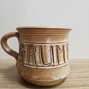 Könnte beinhalten: Eine braune Keramiktasse mit dem Wort "GRUMPY" auf der Seite. Die Tasse hat eine abgerundete Form, einen Henkel und eine gesprenkelte Textur. Die Tasse steht auf einer Holzoberfläche.
