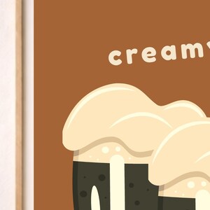 以下が含まれることがあります： 茶色の背景に白い文字で「creamy」と書かれた額装されたウォールアートプリント。イラストには、濃い茶色の液体が入った2つの濃い茶色のグラスが描かれており、その上には側面を滴り落ちる厚い薄ベージュ色の泡が乗っています。