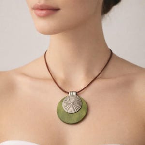 May include: A necklace with a brown cord and a pendant featuring a green wooden disc and a silver spiral design. The pendant is a circular shape. The necklace is worn around the neck.