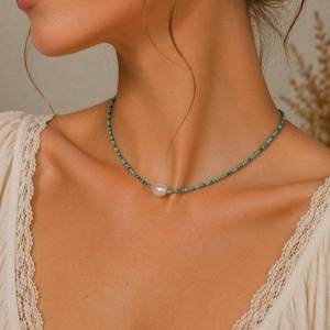 Puede incluir: Un delicado collar con pequeñas cuentas turquesas y detalles dorados. Una sola perla blanca grande es el punto focal del collar. El collar se lleva alrededor del cuello.