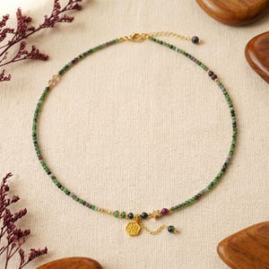 Pode incluir: Um colar delicado com pequenas contas verdes e vermelhas. O colar tem um fecho dourado e um pequeno pingente dourado com caracteres chineses. Uma estrela e uma conta pendurada também estão incluídas no design.