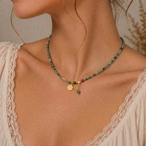 May include: A beaded necklace featuring green, red, and black beads. The necklace has a gold-colored pendant with a small charm and a star-shaped accent. The necklace is worn around the neck.
