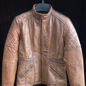 Puede incluir: Chaqueta de cuero marrón claro con diseño acolchado en los hombros. La chaqueta tiene cuello alto, cremallera frontal y dos bolsillos con cremallera. Cuelga de una percha negra sobre un fondo oscuro.