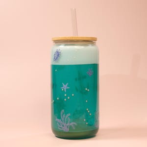 May include: A glass tumbler with a bamboo lid and a clear straw. The tumbler is filled with a teal liquid and decorated with sea-themed illustrations. The design includes a starfish, seashells, and other aquatic elements.