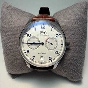 Puede incluir: Un reloj de pulsera automático IWC Schaffhausen plateado con esfera blanca, números negros y correa de cuero marrón. El reloj presenta dos subesferas y el texto "AUTOMATIC" y "SWISS MADE".