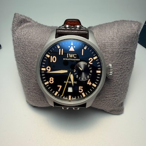 Pode incluir: Um relógio automático IWC Schaffhausen em aço inoxidável com mostrador azul escuro, algarismos e ponteiros luminescentes. O relógio tem uma bracelete de couro castanho e é exibido numa almofada cinzenta. O mostrador mostra a data e o dia.