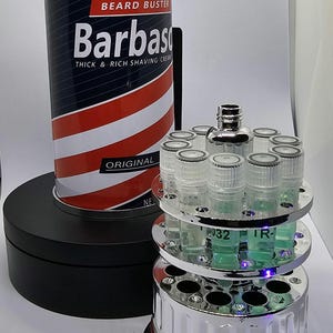 May include: A can of "Barbasol" shaving cream with red, white, and blue stripes, and a chrome-plated device with multiple clear vials containing a green liquid. The shaving cream can is on a black rotating stand.