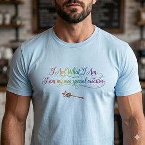 May include: Pale blue t-shirt featuring the text "I Am What I Am, I am my own special creation" in rainbow hues. A star-topped wand is below the text. The shirt has a crew neck and short sleeves.