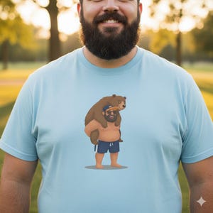 May include: Light blue t-shirt featuring a cartoon illustration of a bear riding on the shoulders of a man wearing sunglasses and blue shorts. The bear is brown, and the man has a beard. The shirt is a solid color.