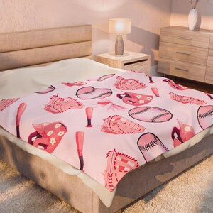 Op de afbeelding: Een roze baseball-thema deken met illustraties van honkballen, handschoenen en knuppels. Het ontwerp bevat verschillende tinten roze en rood, wat een speelse en sportieve uitstraling creëert. De deken ligt op een bed.