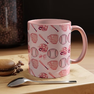 Puede incluir: Una taza de cerámica rosa con un diseño de béisbol. El diseño incluye pelotas de béisbol, bates, guantes y cascos en tonos rosas. La taza tiene un asa y un interior rosas, perfecta para café o té.