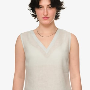 Könnte beinhalten: Ein ärmelloses, hellblaues Oberteil mit V-Ausschnitt. Das Oberteil ist aus leichtem Stoff gefertigt und wird mit einer zarten Silberkette kombiniert. Das Model hat kurze, dunkle Haare und kleine Creolen. Der Stil ist minimalistisch und lässig.