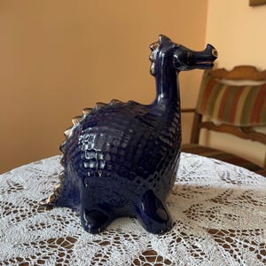 May include: A ceramic dragon figurine in a deep blue glaze with gold accents. The dragon has a textured body, a long neck, and a detailed head. The figurine is displayed on a white lace tablecloth.