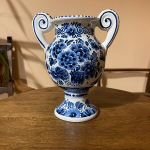 May include: A white ceramic vase featuring intricate blue floral patterns and scrollwork. The vessel has a wide, flared rim, a bulbous body, and two prominent, curled handles on either side. The base is pedestal-style with matching blue floral motifs.