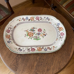 May include: An oval ceramic serving platter with a cream-colored base featuring a central illustration of a small bird perched on a flowering tree. The rim is decorated with a repeating floral pattern in shades of orange, red, blue, and green.