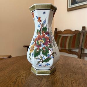 May include: A white ceramic vase with a hexagonal base and rim, adorned with a colorful floral design. The flowers are in shades of red, orange, yellow, and purple, with green leaves and blue accents. The vase is on a wooden table.