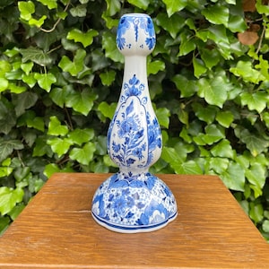 May include: A white ceramic candlestick holder featuring a bulbous central stem and a wide circular base, decorated with intricate blue floral patterns and scrolling leaf motifs painted in a traditional style across the entire exterior surface.