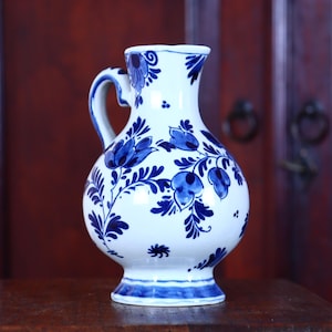 May include: White ceramic pitcher with a blue floral design. The pitcher has a rounded body, a handle, and a flared base. The blue floral pattern includes stylized flowers and leaves. The pitcher is likely for decorative use or serving beverages.