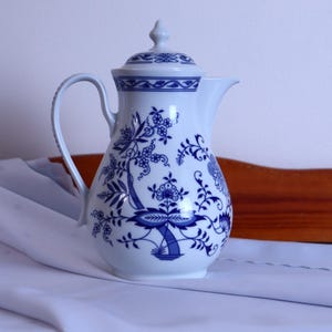 May include: A white porcelain coffee pot featuring a blue onion pattern with intricate floral and botanical motifs. The vessel includes a curved handle, a pointed spout, and a matching lid with a small finial, all decorated in blue.