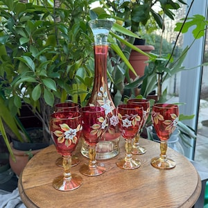 May include: A tall glass decanter and four matching stemmed glasses, all featuring a deep red hue with hand-painted white floral motifs and green leaves. The decanter has a clear glass stopper, and each piece rests on a clear, flared pedestal base.