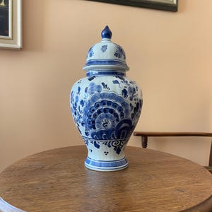 May include: A white ceramic ginger jar with a removable lid, featuring intricate hand-painted blue floral and foliate patterns. The vessel has a bulbous body, a tapered neck, and a pointed finial on the lid, decorated with blue bands and dots.