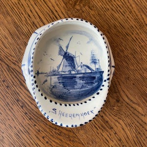 May include: A white ceramic bowl with a blue hand-painted illustration of a windmill and a house near water. The rim features a dotted blue pattern, and the text 'S. HEERENHOEK' is written in blue on the front edge of the bowl's interior surface.