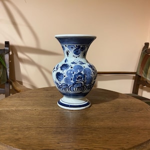 May include: A ceramic vase with a white base and blue hand-painted floral patterns. The vase features a flared rim, a rounded body, and a pedestal base. The blue designs include various flower motifs and horizontal bands around the neck and base.