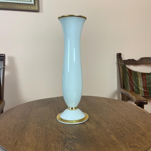 May include: A tall, light blue vase with a flared top and a pedestal base. The vase has gold trim around the top and base. The vase is sitting on a round, wooden table.
