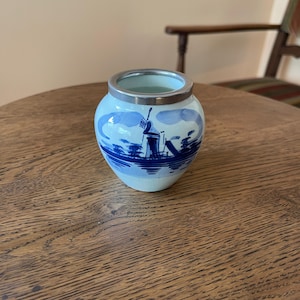 May include: A small, round ceramic vase with a light blue base and a silver rim. The vase features a blue and white windmill scene. The design includes a windmill, water, and clouds. The vase is sitting on a wooden table.