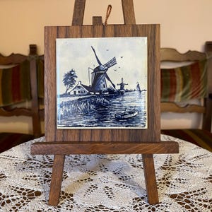 May include: A framed ceramic tile featuring a blue and white windmill scene, displayed on a small wooden easel. The artwork depicts windmills, a boat, and a waterside landscape. The frame and easel are made of dark wood.