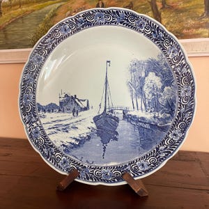 May include: A white ceramic plate with a blue and white landscape scene. The plate features a sailboat on a canal, a house, and trees. The rim has a blue floral design. The plate is displayed on a wooden stand.