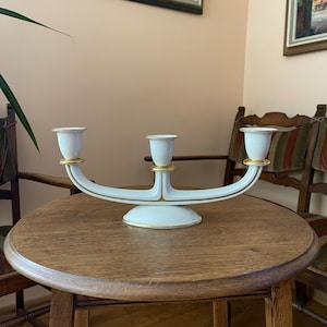 May include: A white porcelain candelabra with three candle holders, featuring a curved base and gold trim accents on the rims and along the central structure. The piece has a smooth, glossy finish and a symmetrical design with a central pedestal.