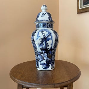 May include: A white ceramic ginger jar with a removable lid, featuring blue hand-painted illustrations of a traditional windmill in a landscape setting, surrounded by intricate floral and scrollwork patterns along the neck and base.