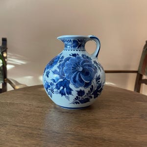 May include: A round ceramic pitcher with a white base and blue floral patterns. The vessel features a wide, bulbous body, a short neck, a flared rim, and a single curved handle. Intricate blue painted flowers and leaves decorate the exterior.