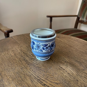 May include: A ceramic cup featuring a white base with blue floral patterns and horizontal bands. The cup has a rounded body and a smooth, metallic silver-colored rim at the top. The interior of the cup is white, and the exterior displays intricate blue painted flowers and leaves.