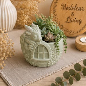 May include: A light green ceramic planter shaped like a small house with a sleeping dragon figure resting on the roof. The planter is filled with various green succulents, including a trailing string of pearls plant, and features a window design.