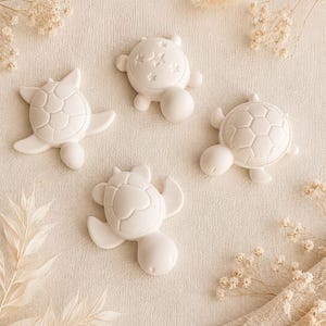 Peut inclure: Quatre ornements en forme de tortue en plâtre ou en argile blanc mat. Chaque tortue présente un motif de carapace distinct, incluant des hexagones géométriques, une forme de cœur et un motif d'étoiles. Les figurines sont disposées en groupe.