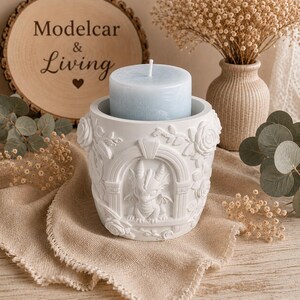 May include: A white cylindrical candle holder featuring a relief sculpture of a small dragon framed by an archway and surrounded by rose blossoms. A light blue pillar candle sits inside the holder, which is crafted from a matte white material.