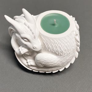 Può includere: Un portacandele bianco a forma di drago raggomitolato con squame e ali testurizzate. L'apertura circolare contiene una candela tea light rotonda verde chiaro con un piccolo stoppino bianco centrato sulla superficie della cera.