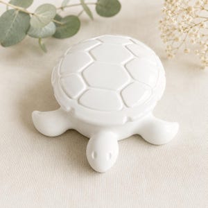 Könnte beinhalten: Eine weiße, schildkrötenförmige Schmuckdose aus Keramik mit glatter, glänzender Oberfläche. Der Deckel ist mit einem geformten, sechseckigen Panzermuster versehen, während die Basis die Form von Kopf und Flossen einer Schildkröte aufweist.