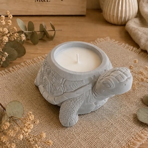 May include: A light grey concrete candle holder shaped like a turtle with a textured shell and head. The centre of the turtle shell features a circular opening containing a white wax candle with a single centred cotton wick standing upright.