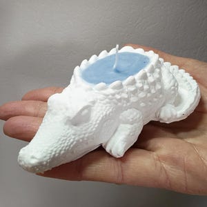 May include: A white alligator-shaped candle with a light blue wax interior. The candle has a textured surface and a wick. The alligator is lying down, with its head and tail visible.