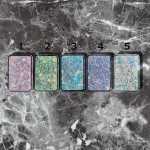 May include: Five rectangular cosmetic compacts with glitter in various colors: purple, green, blue, lavender, and teal. Each compact has a clear plastic case. The glitter is densely packed, creating a sparkling effect. The compacts are arranged in a row on a marble surface.