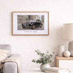 May include: Framed print of a black Citroen 11BL 1938/39 car with yellow wheels, parked on a city street. The artwork is in a wooden frame and hangs on a white wall. The print includes the car's model and year.