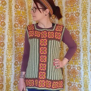 May include: A handmade crochet vest in shades of green, brown, and pink, featuring a square neckline and a patterned design. The vest is paired with a matching headband. The background is a vintage floral wallpaper.